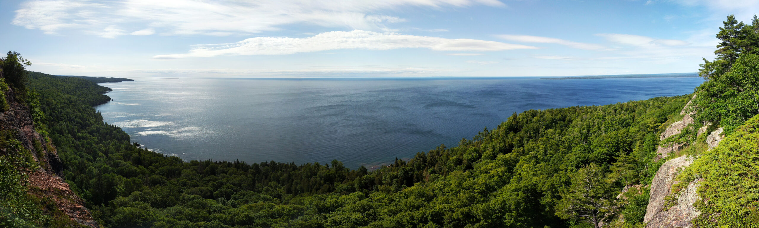 A panoramic photo overlooking a large lake from a tall vantage point. The foreground of the image is filled with dense green forest, while the background is dominated by a large, calm, blue lake. The sky is bright blue and mottled with white clouds.